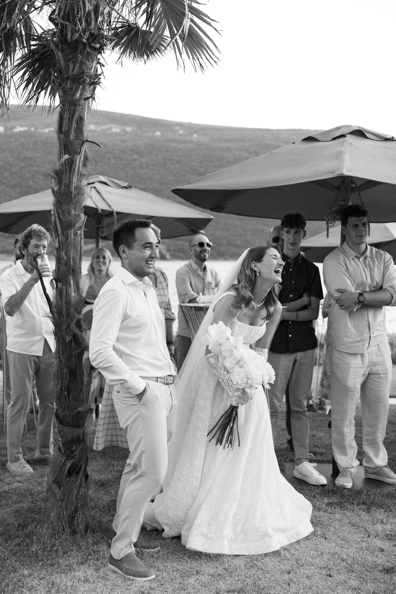 A black and white wedding photo outdoors with a couple in white attire sharing a candid moment under umbrellas and palm trees.