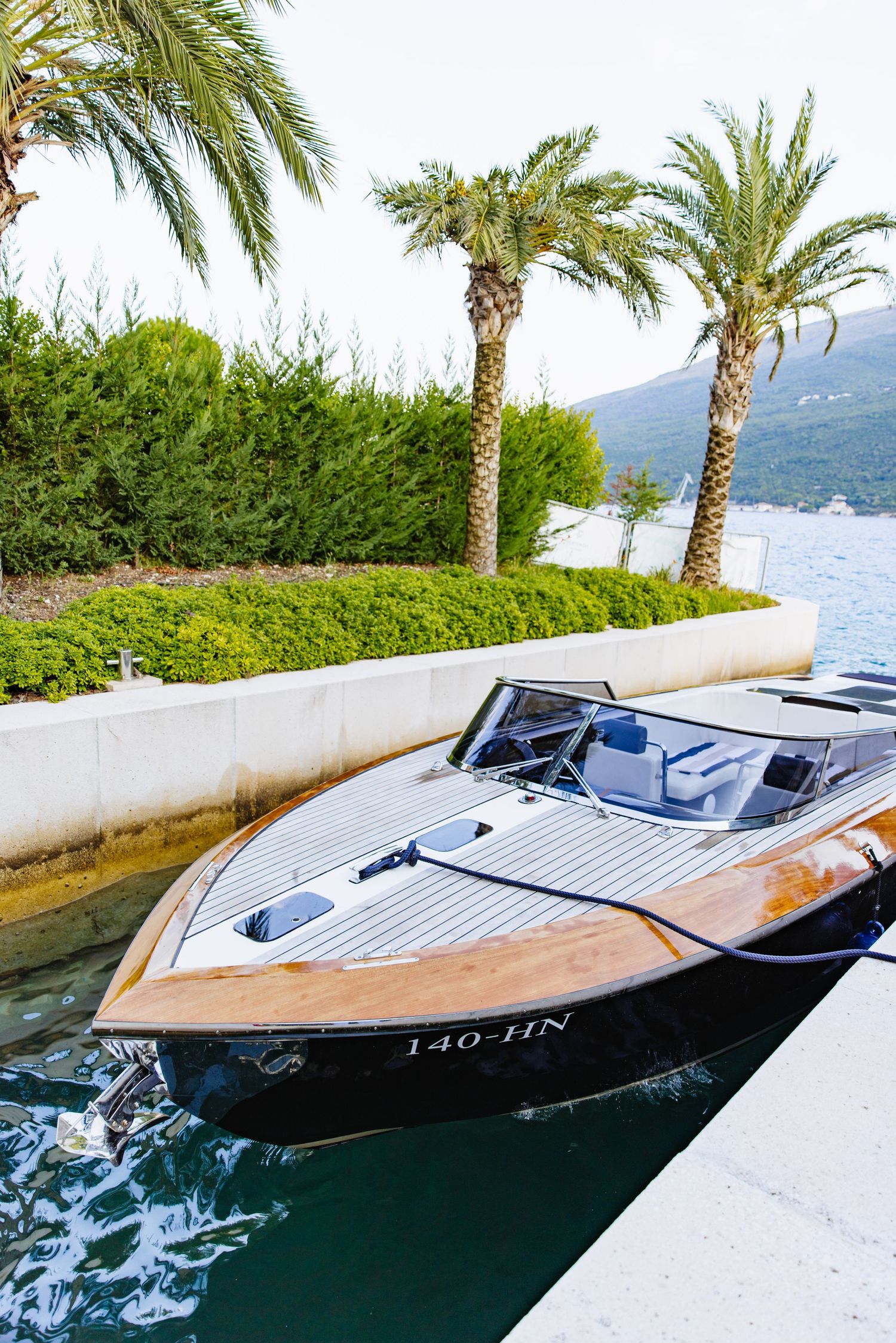 Luxury wooden speedboat moored at Mediterranean dock with palm trees and mountains in background.