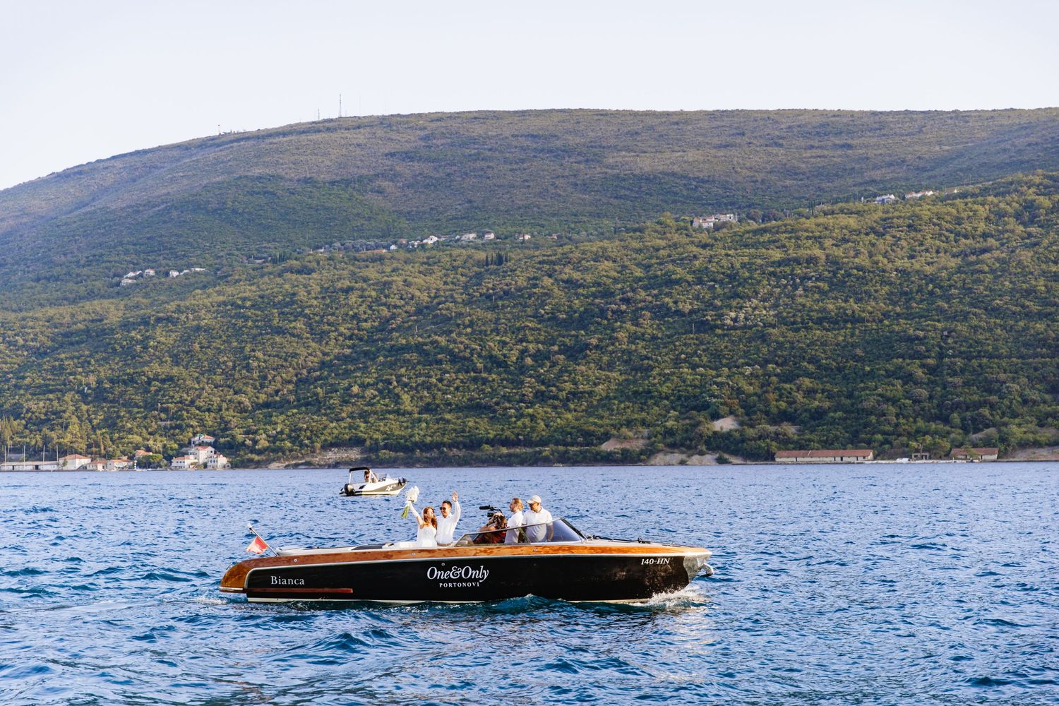 A classic wooden boat cruises on blue waters near a green mountainous shoreline on a sunny day.
