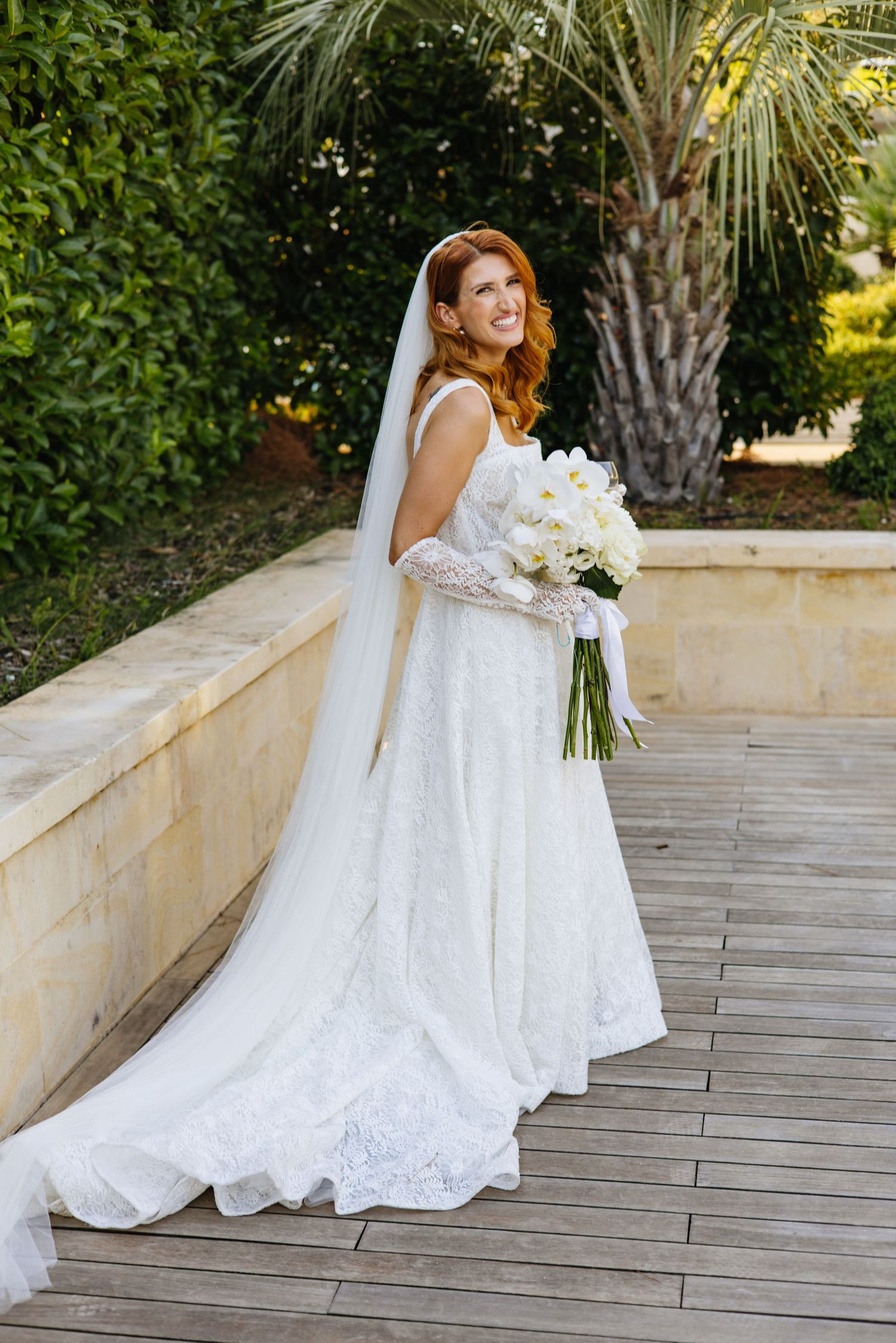 A bride in a elegant white lace wedding dress holds a bouquet of white flowers on a wooden deck with palm trees.