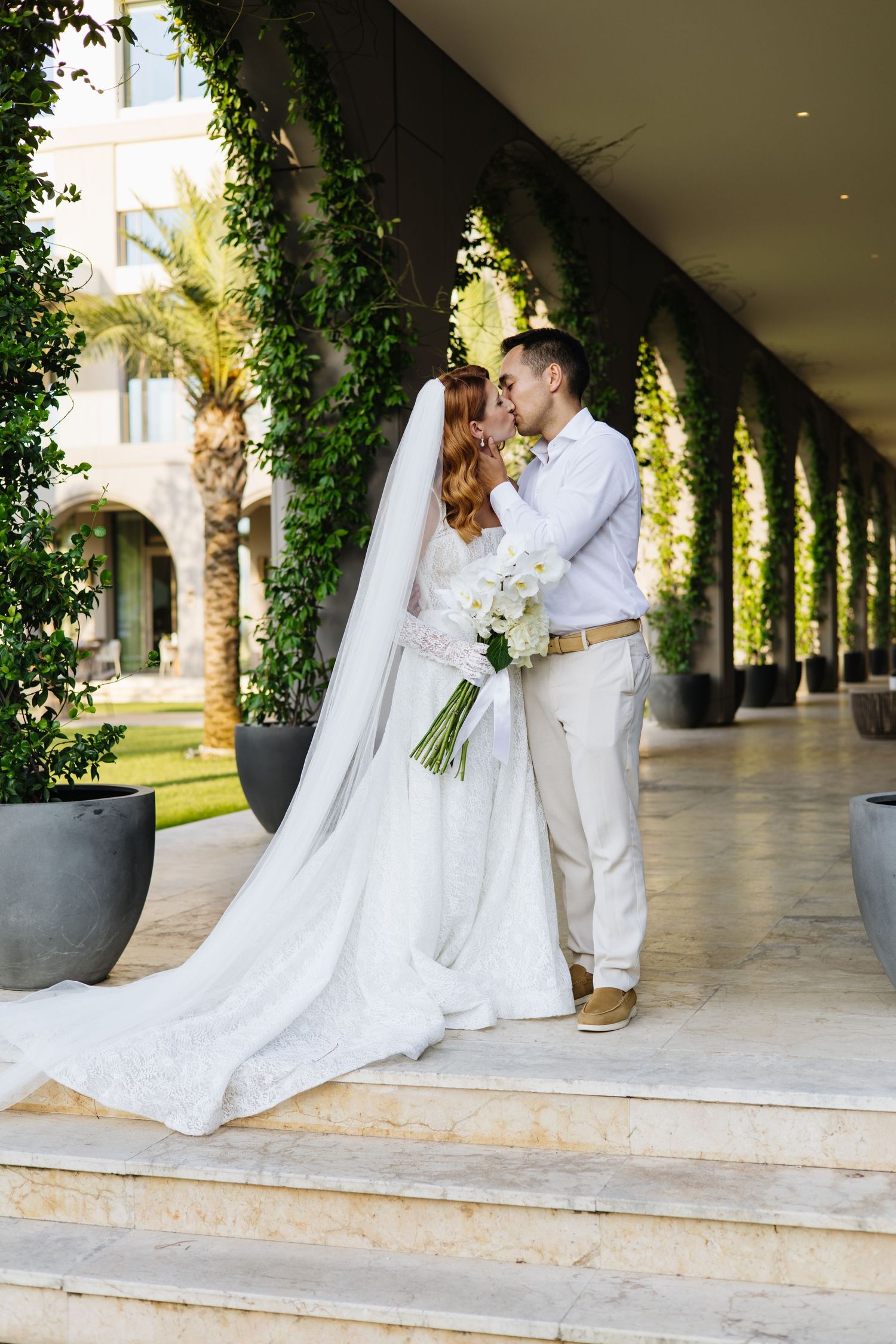 A couple in wedding attire shares a romantic moment under a vine-covered colonnade at an elegant venue.
