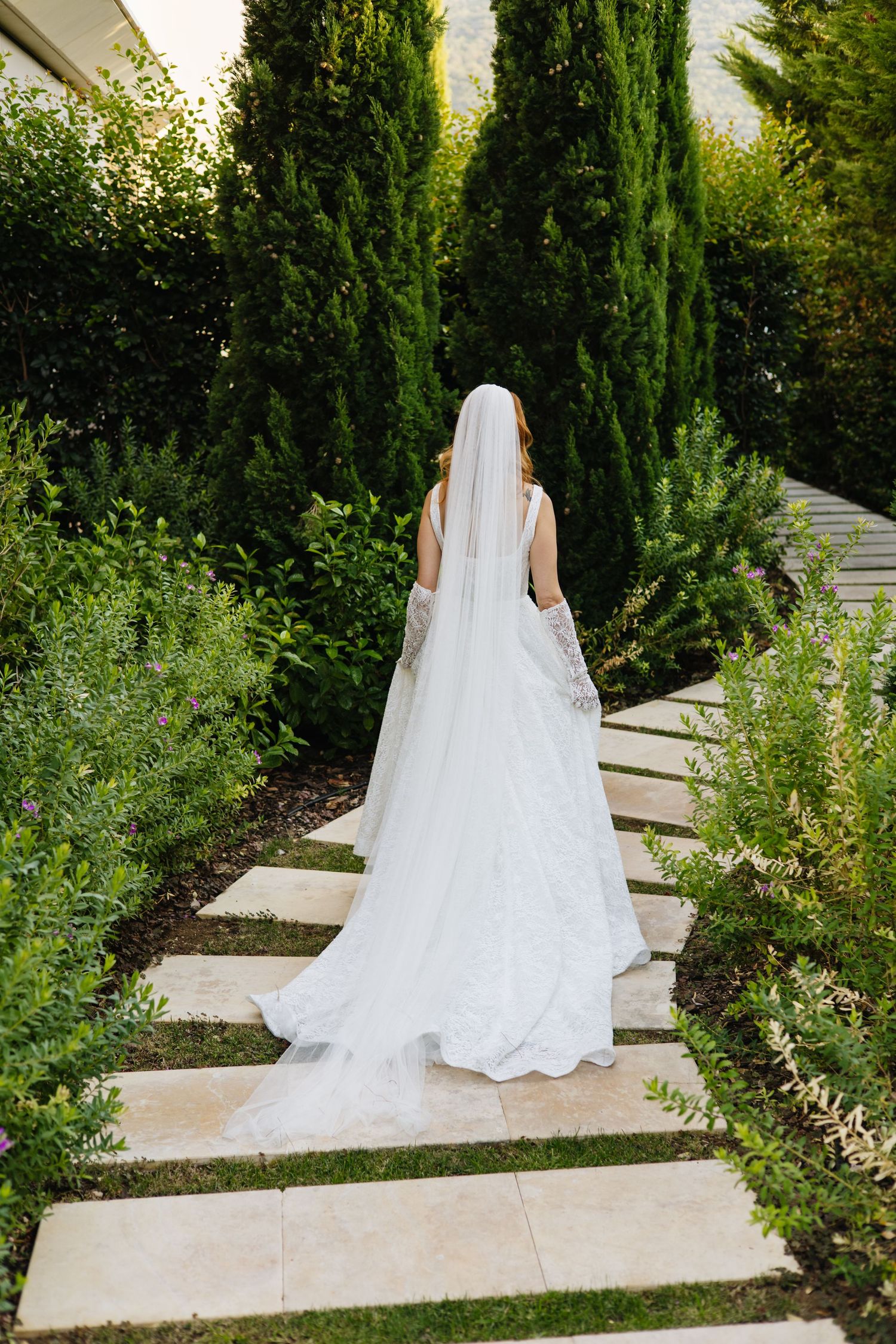 A bride in a white lace wedding gown with long veil walks up stone garden steps flanked by cypress trees and greenery.