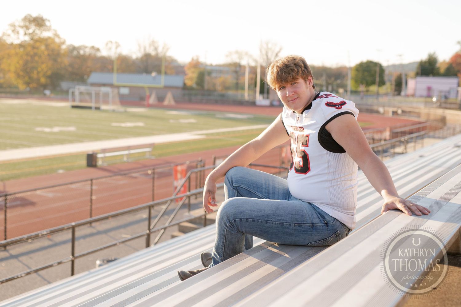 Student athlete sits on metal bleachers wearing white Eagles football jersey during outdoor photo session.