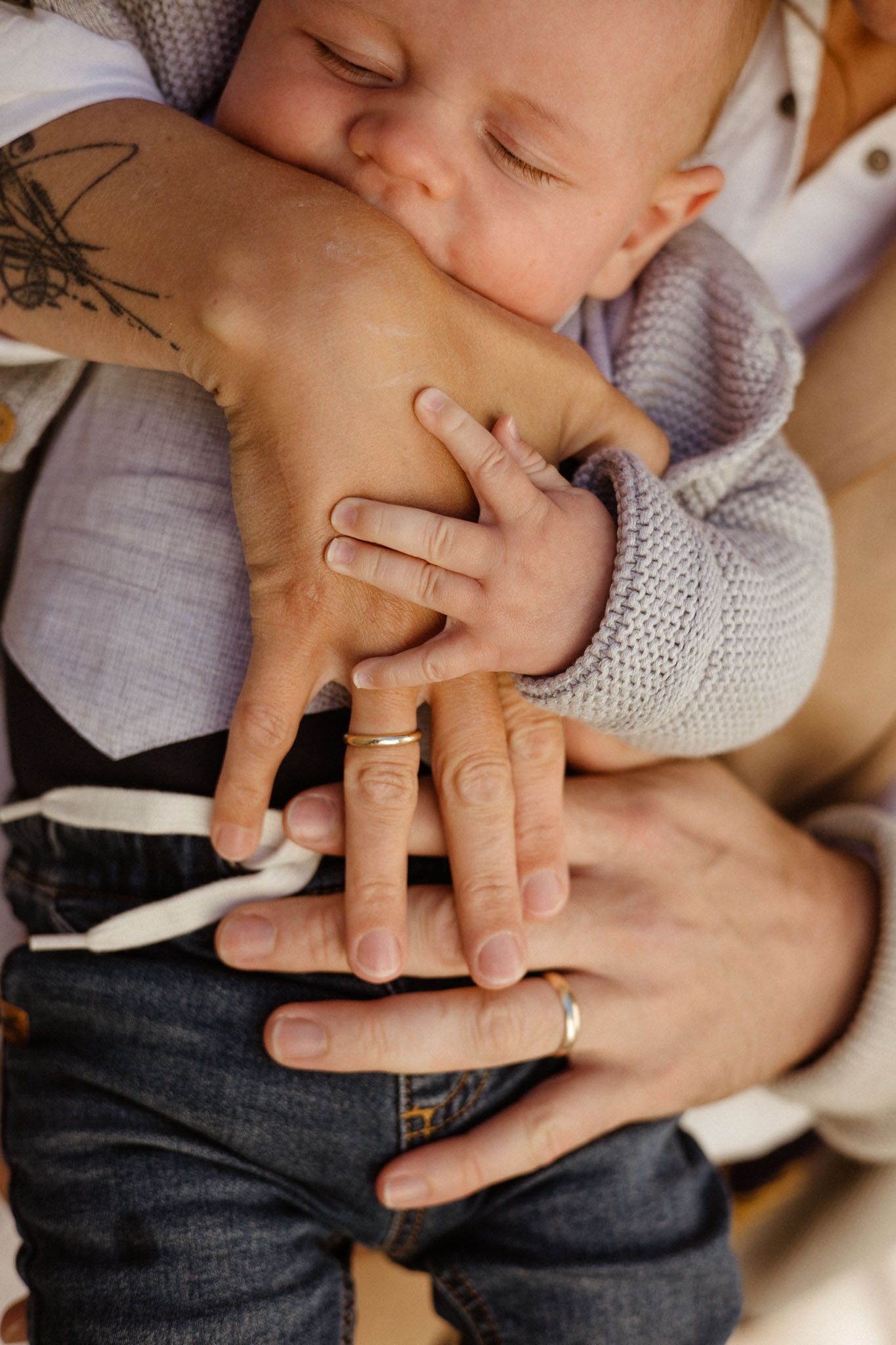 Small baby in gray knit sweater holds parent's tattooed hand with wedding ring in tender moment.