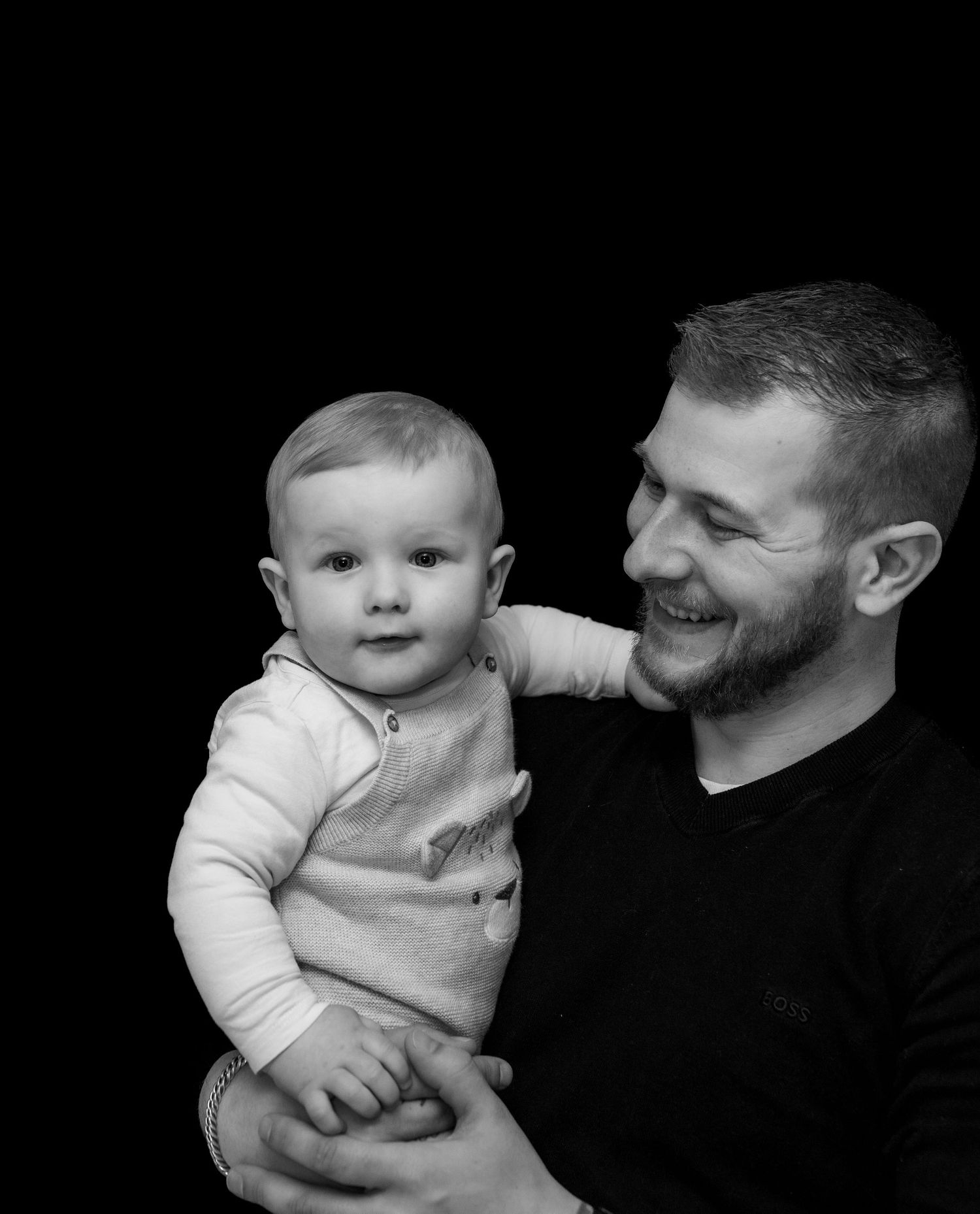 "Lachende vader houdt zijn baby vast, terwijl de baby nieuwsgierig in de camera kijkt. Zwart-wit portret met een warme, liefdevolle sfeer."