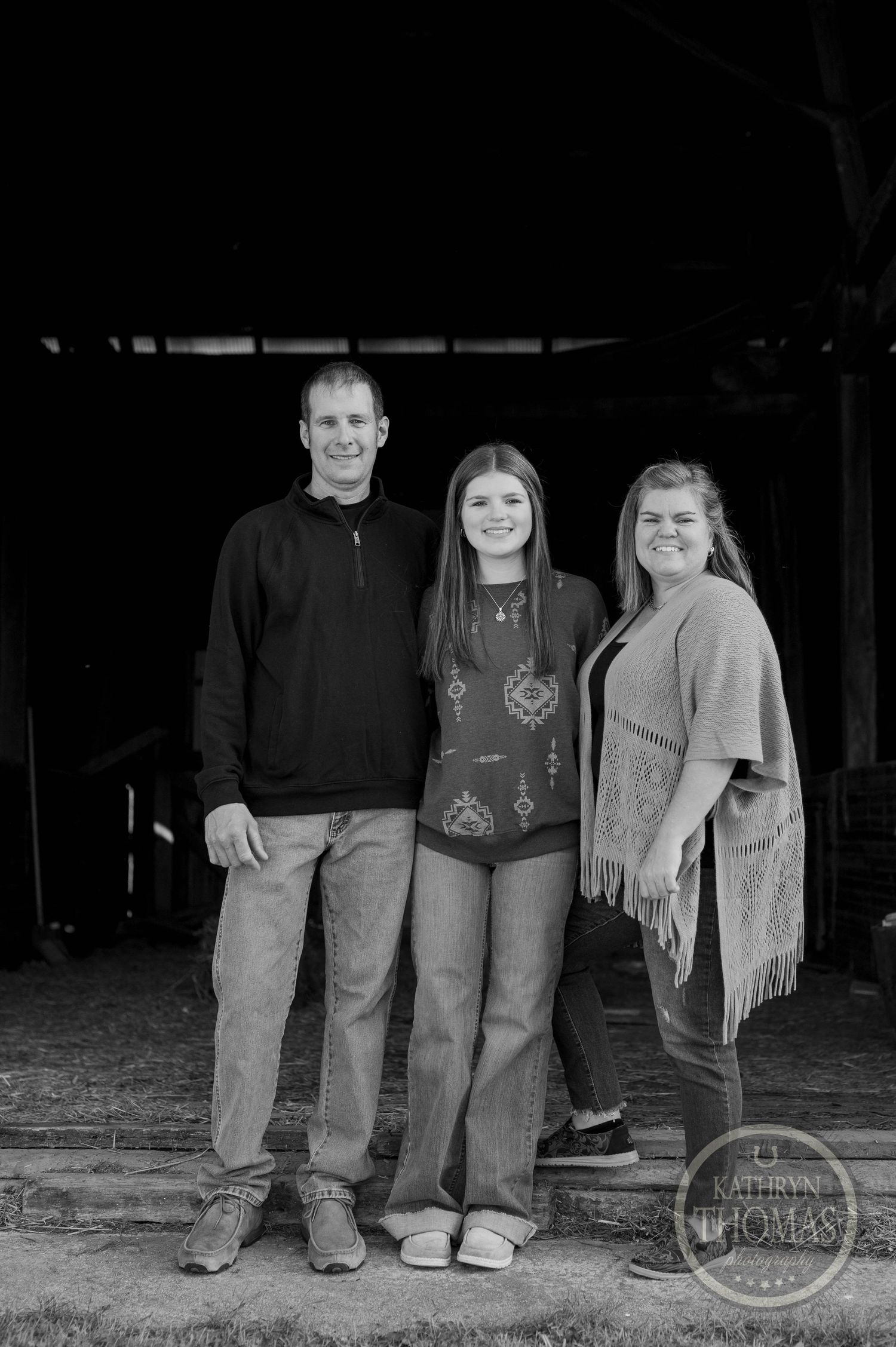 Black and white family portrait of three people standing together in a barn doorway.