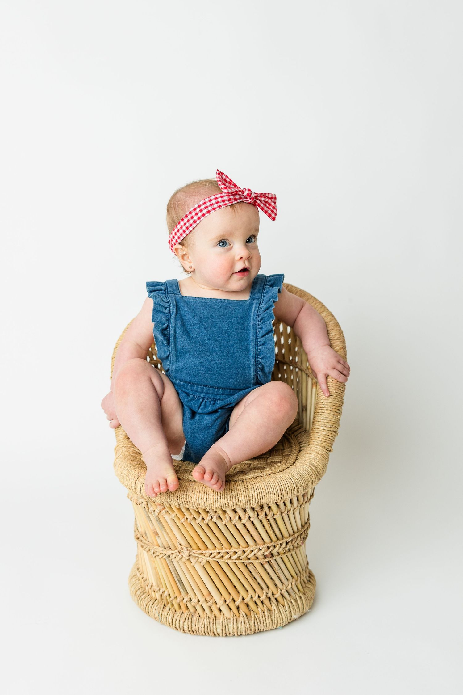Baby wearing blue romper and red gingham headband sits in woven rattan peacock chair against white background.