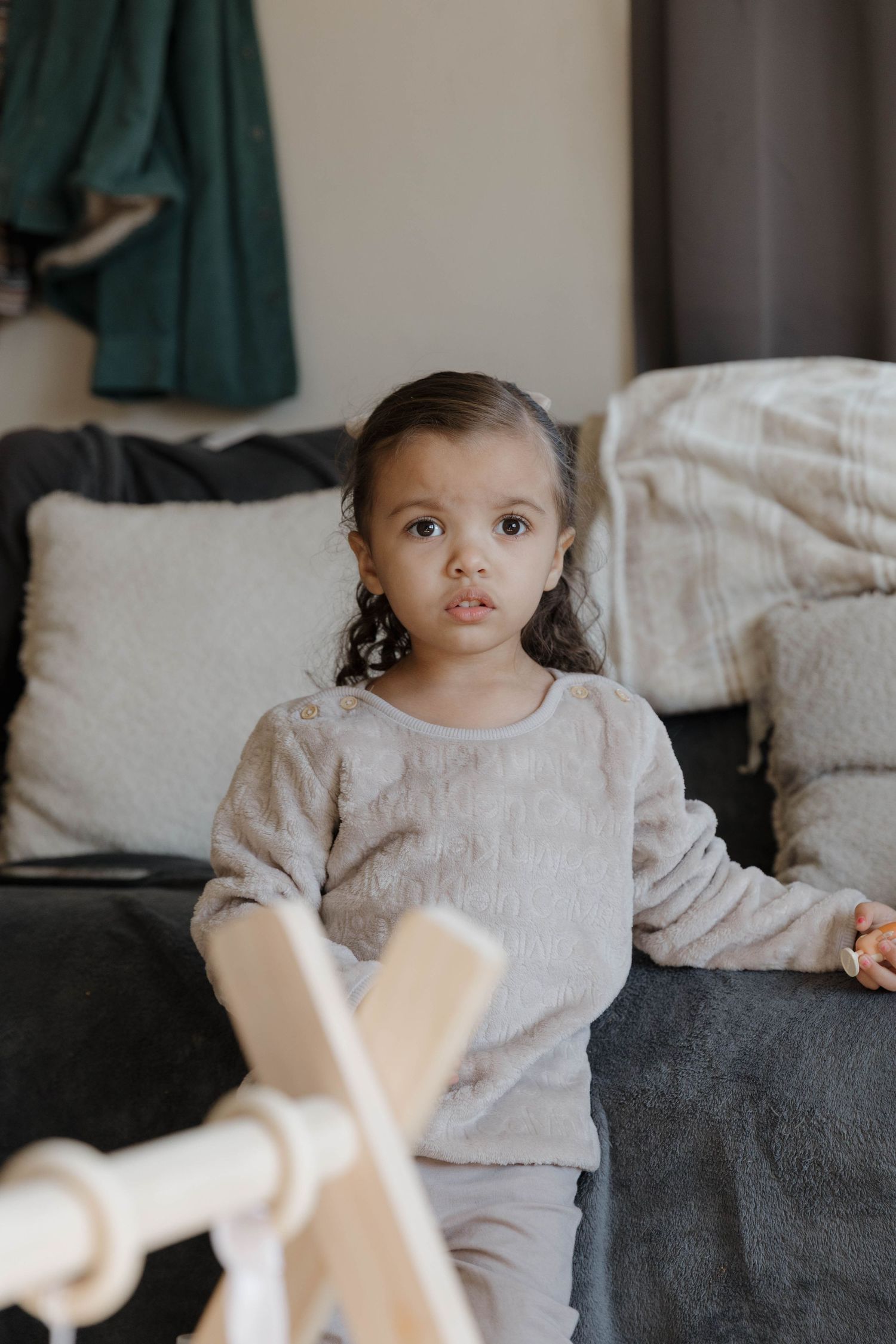 Small child in a light gray sweater sits on a gray couch with soft pillows in a cozy living room setting.