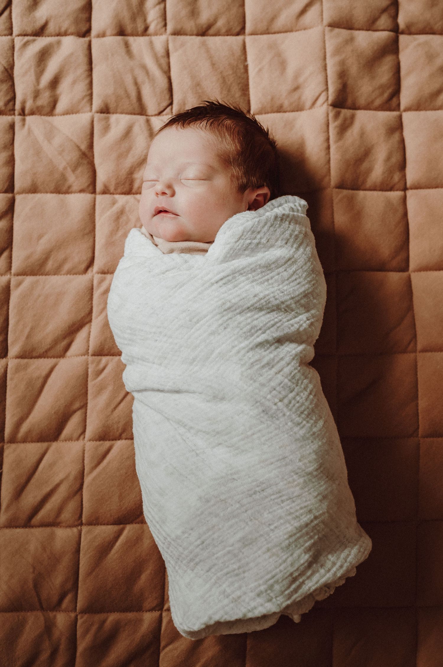 Sleeping newborn swaddled in white blanket on brown quilted background.