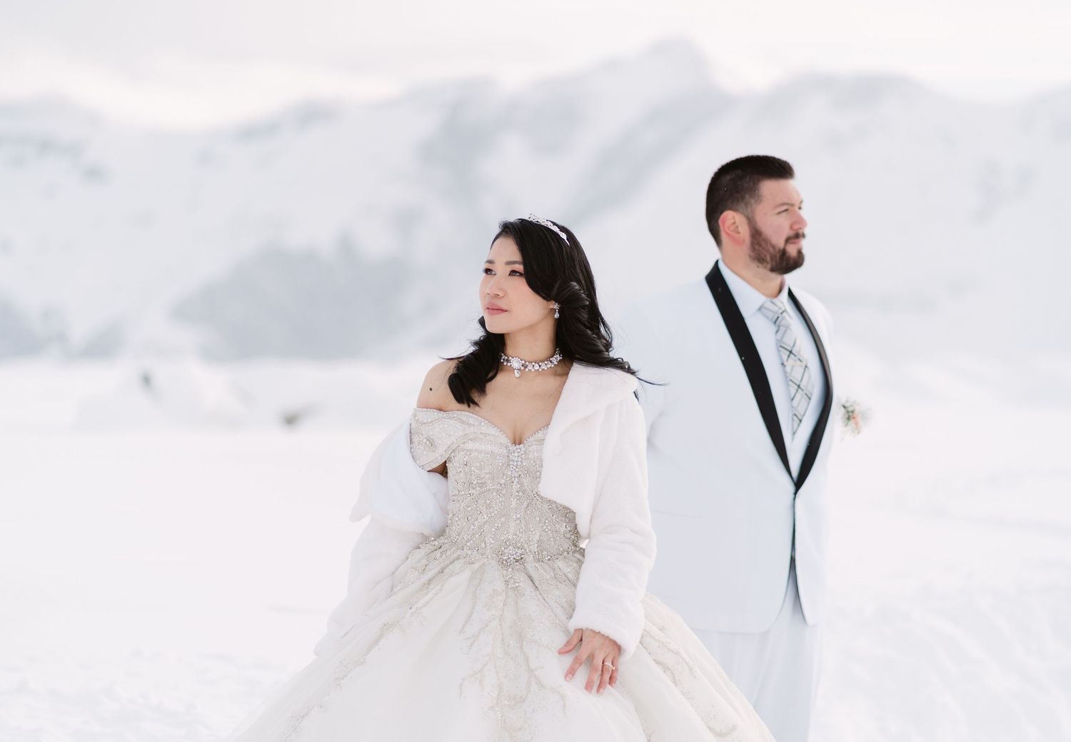 A couple in wedding attire stand together against a snowy winter mountain landscape on their special day.