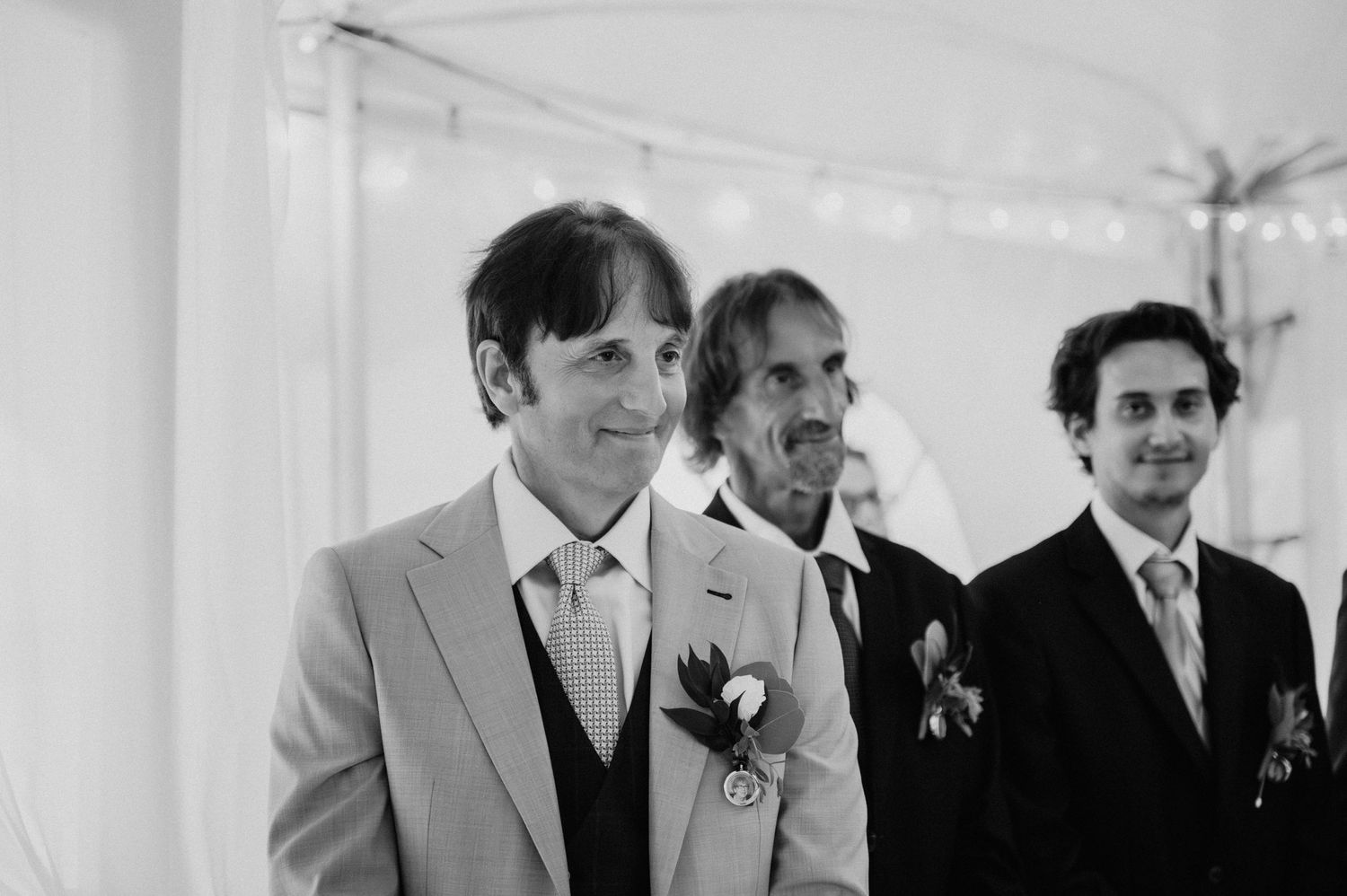 Groomsmen in formal attire stand together under a white tent during wedding festivities.