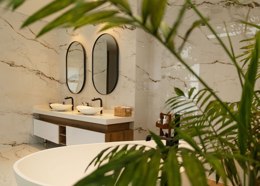 Modern bathroom with marble walls, dual oval mirrors, vessel sinks, wooden vanity, and lush tropical plants in foreground.