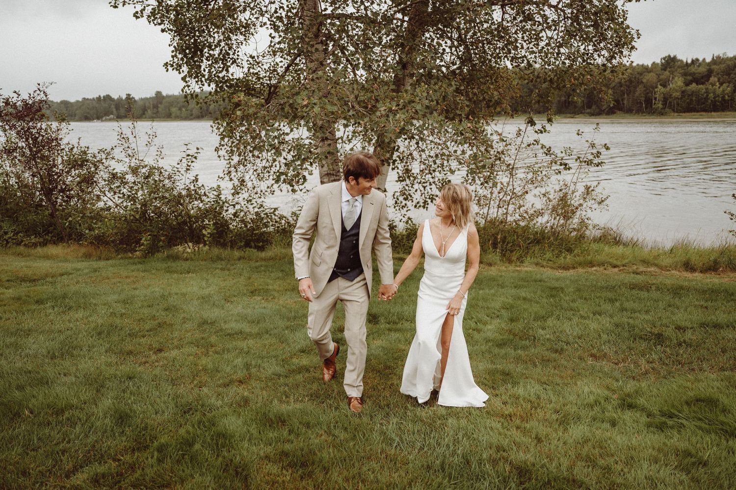 A romantic couple walks together on a grassy lakeside bank during their wedding day photoshoot.