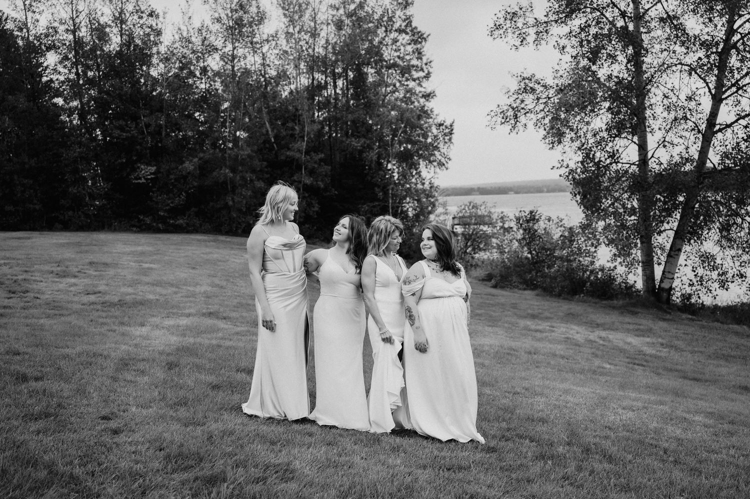 Wedding party in light-colored dresses poses together on grassy hillside near trees.