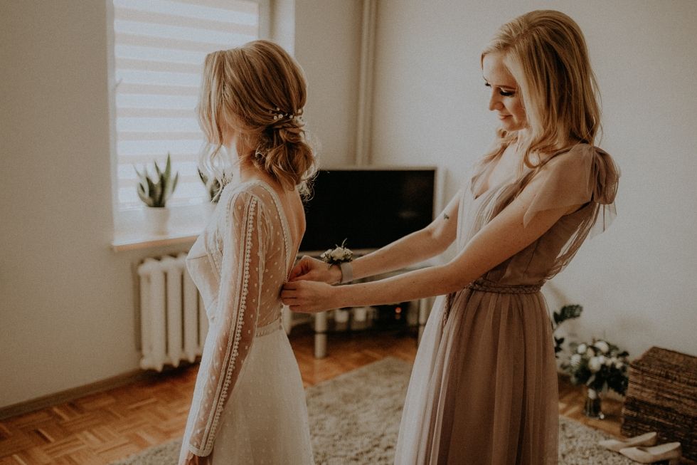 Warm-toned photo of two women in elegant dresses holding hands in sunlit room.