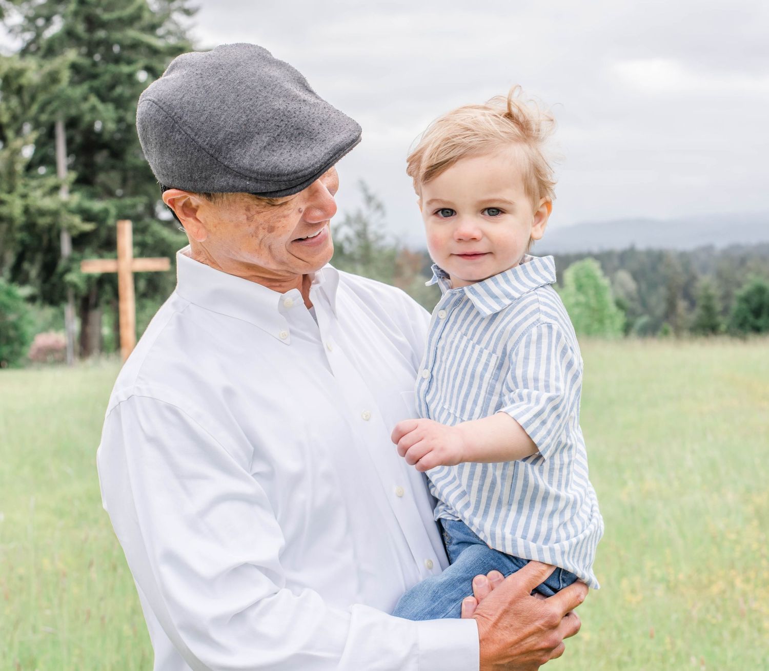 Father and young child share playful moments in a sunlit grassy meadow with evergreen trees in background.