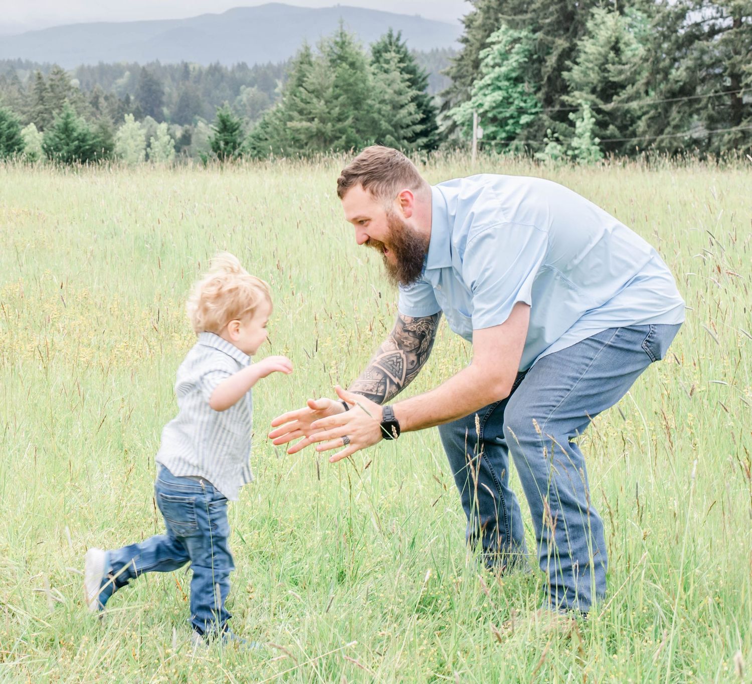 Adult and child playing together in tall grass field with evergreen forest backdrop.