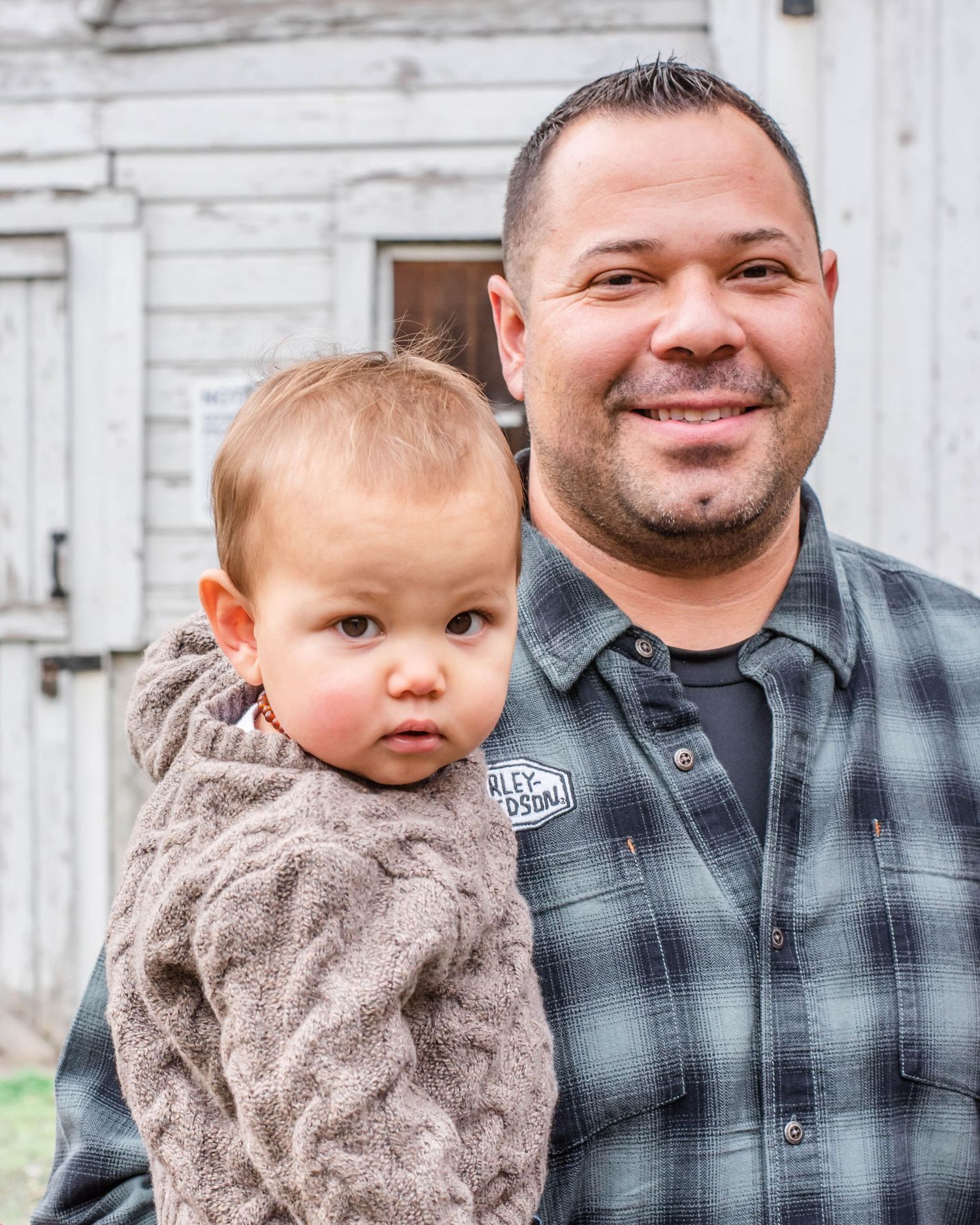 Parent and child wearing casual attire pose together against white wooden building background.