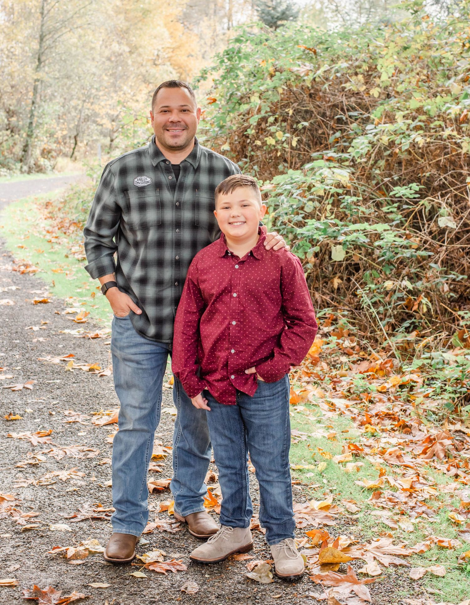 Two people in burgundy and plaid shirts share a candid moment on a leaf-covered path.