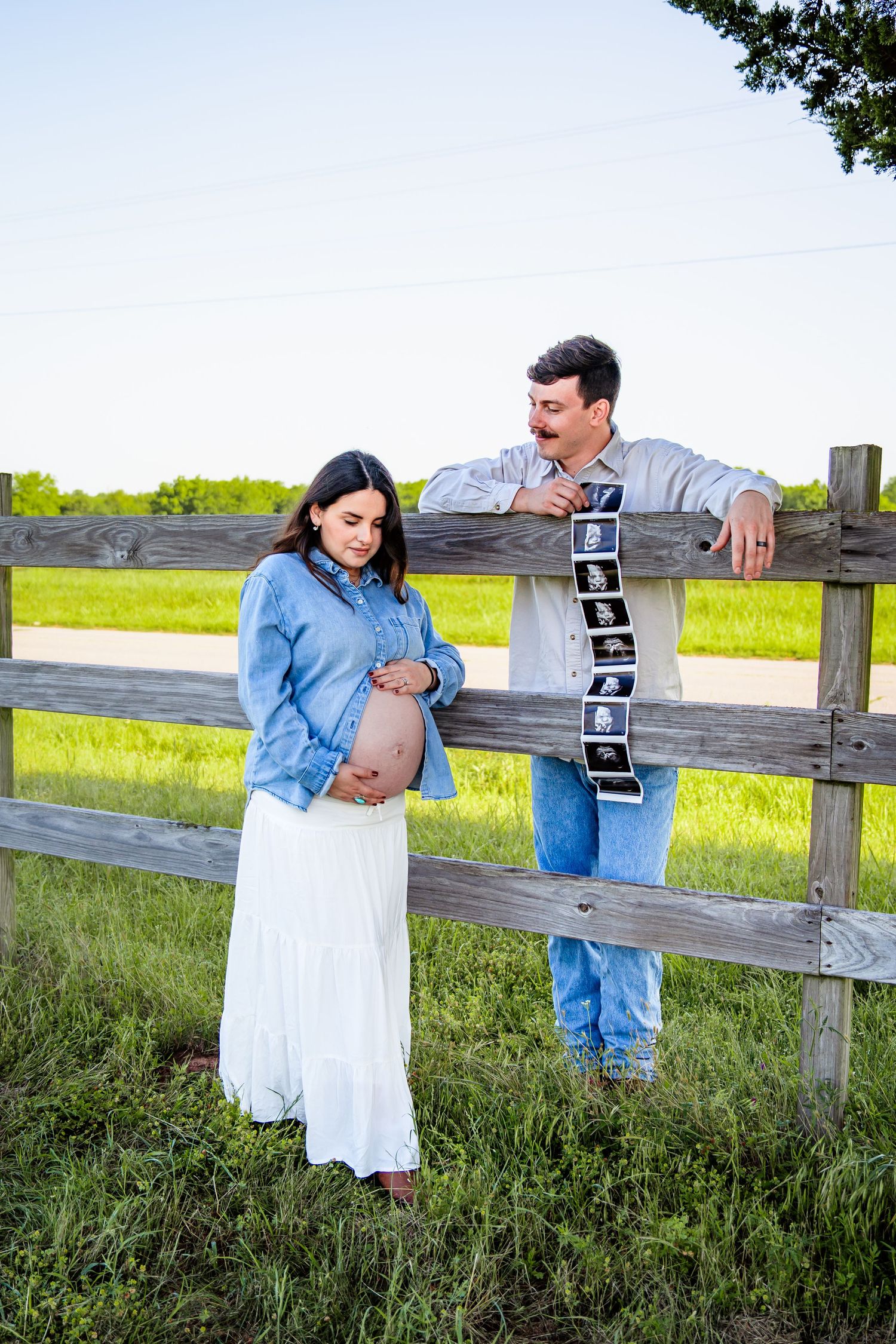 Couple poses with ultrasound photos by a rustic wooden fence in a grassy field.