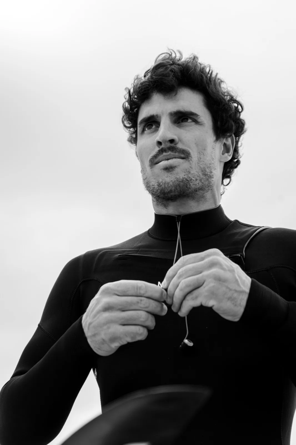 Surfeur debout, tenant sa combinaison au niveau du col, regardant l’horizon avec intensité.