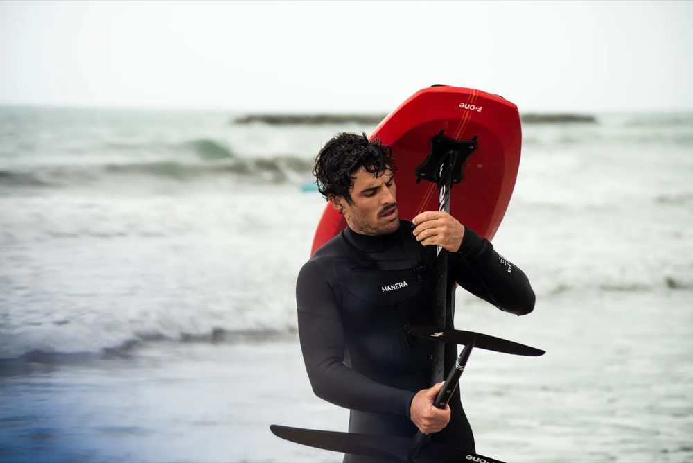 Surfeur sortant de l’eau, portant une planche rouge sur l’épaule, en combinaison noire.