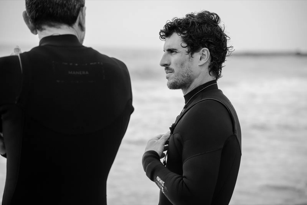 Deux surfeurs en combinaison néoprène discutent au bord de la plage, avec la mer en arrière-plan.