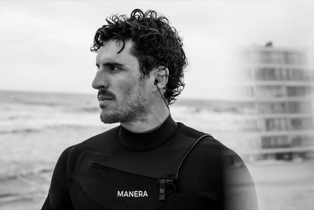 Portrait en noir et blanc d’un homme en combinaison de surf “MANERA”, sur une plage avec un immeuble flouté au second plan.