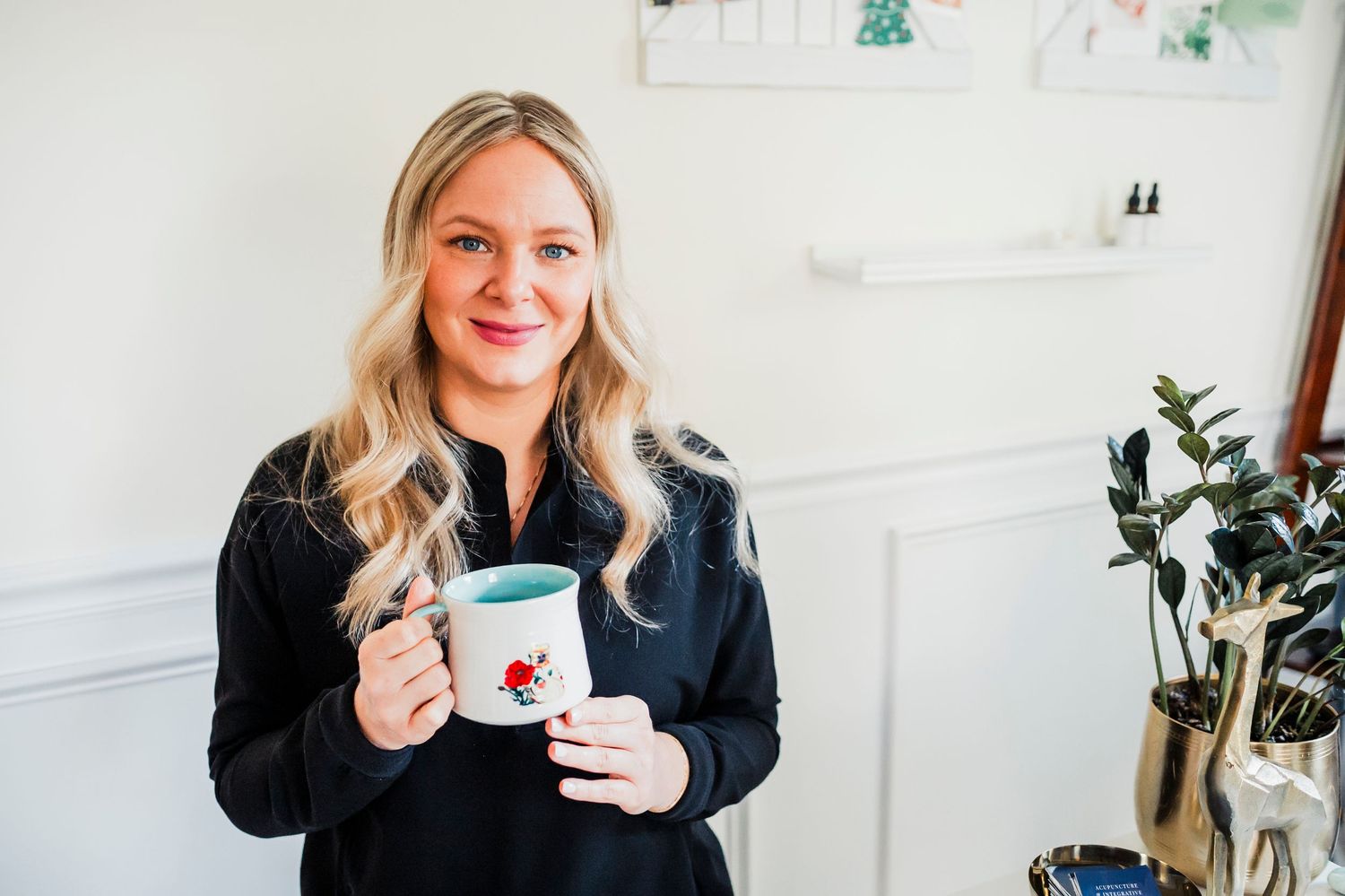 Woman in black sweater holds white ceramic mug while standing against white wall with indoor plants.