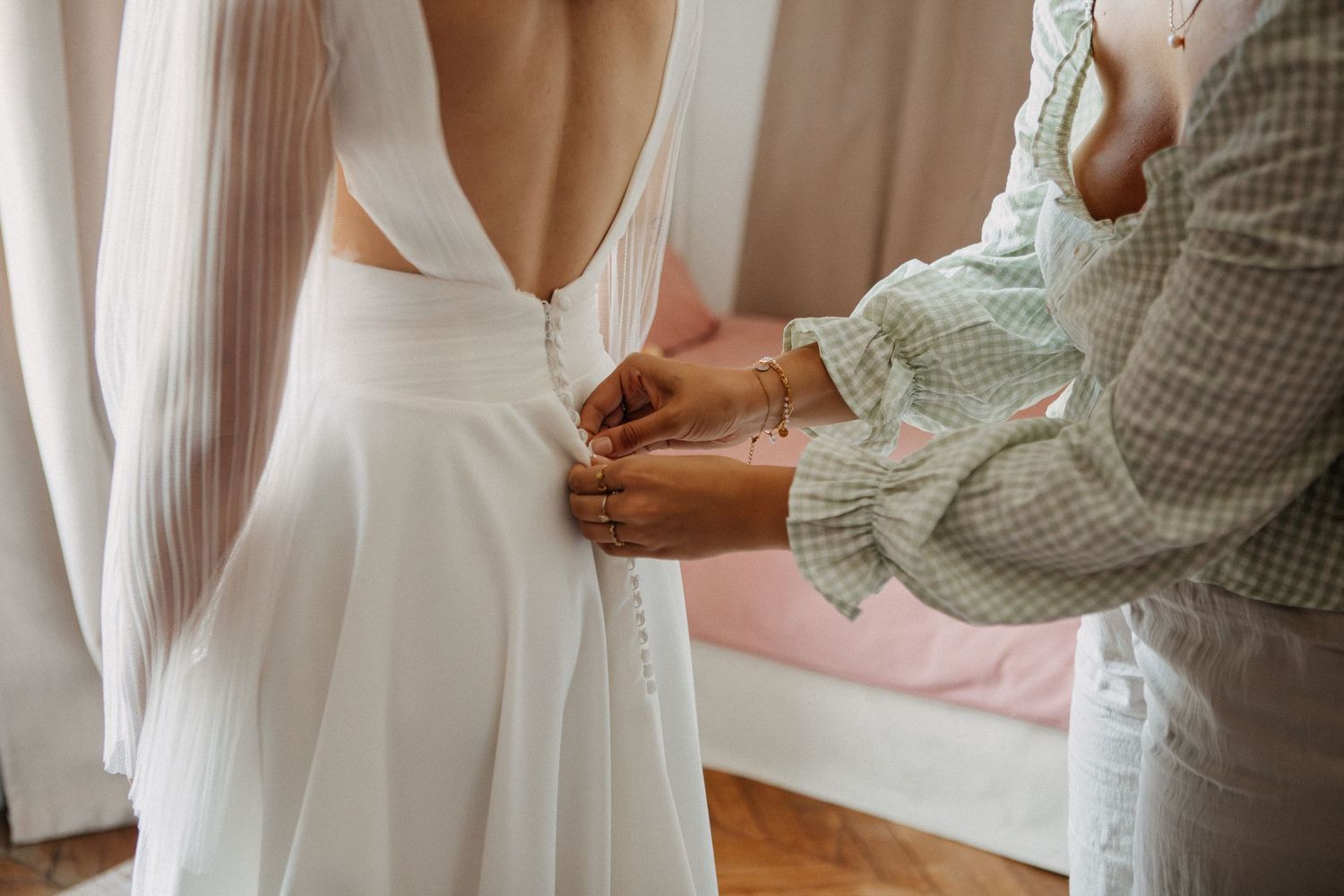 Helper fastening small buttons on the back of a bride's elegant white deep-V wedding dress.