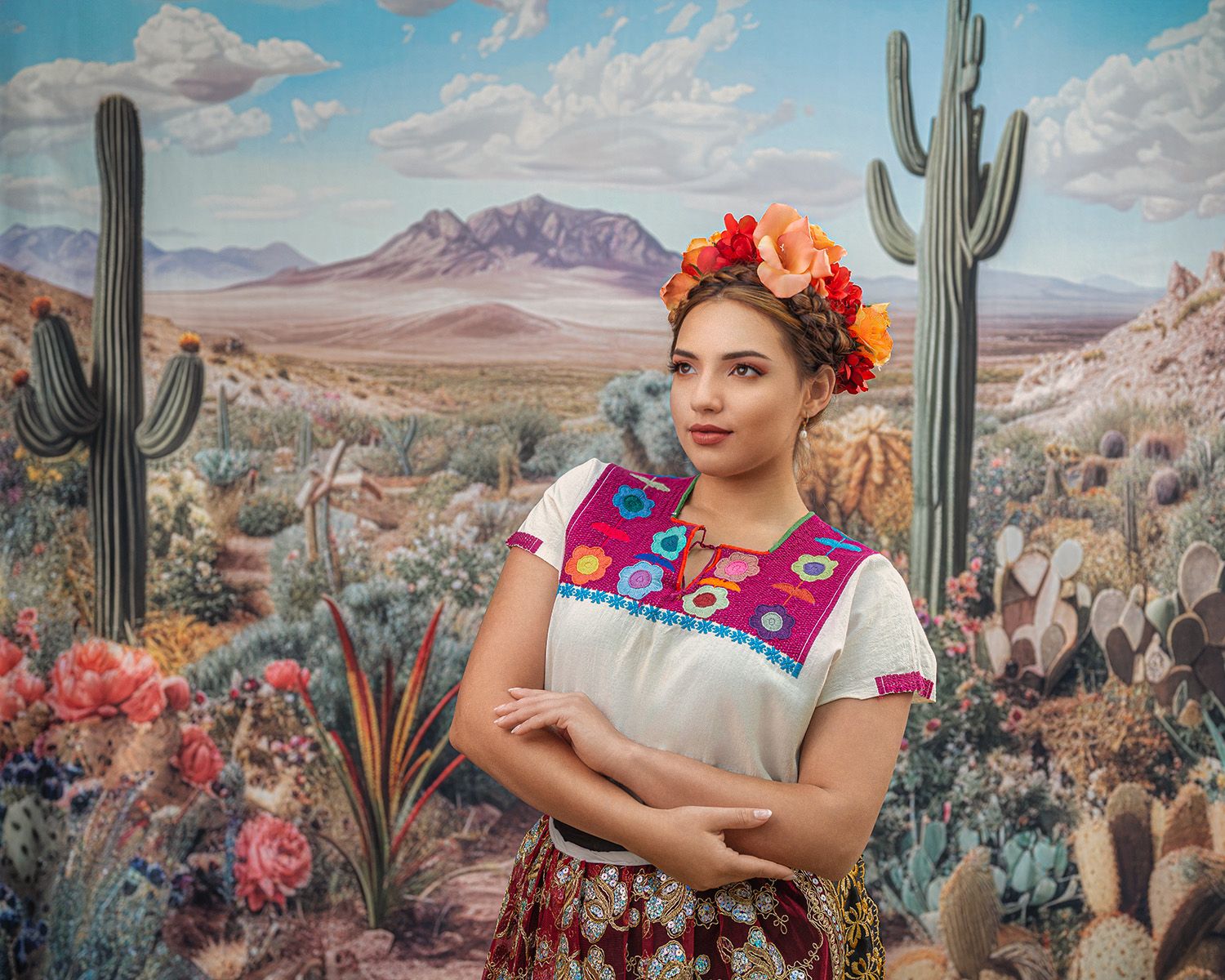 Woman in colorful traditional dress and floral crown stands in front of a desert cactus mural. Photo by Carolina Waldroff.