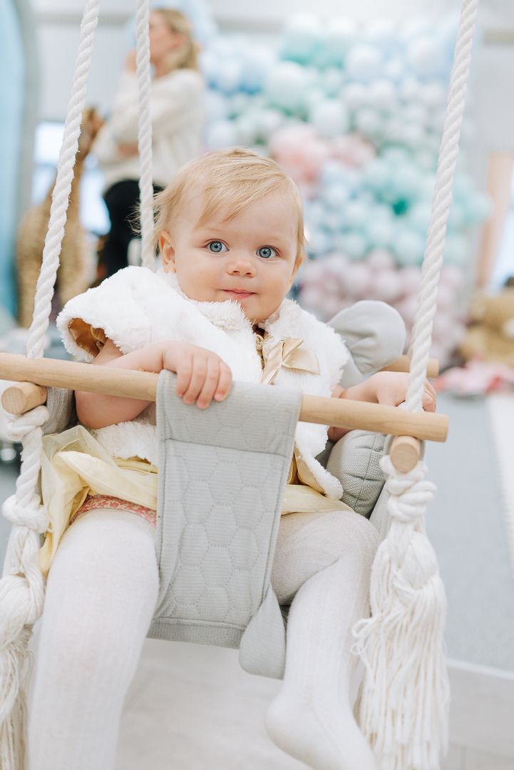 Small child sits bundled in warm winter white clothing on a decorative wooden swing with rope sides.