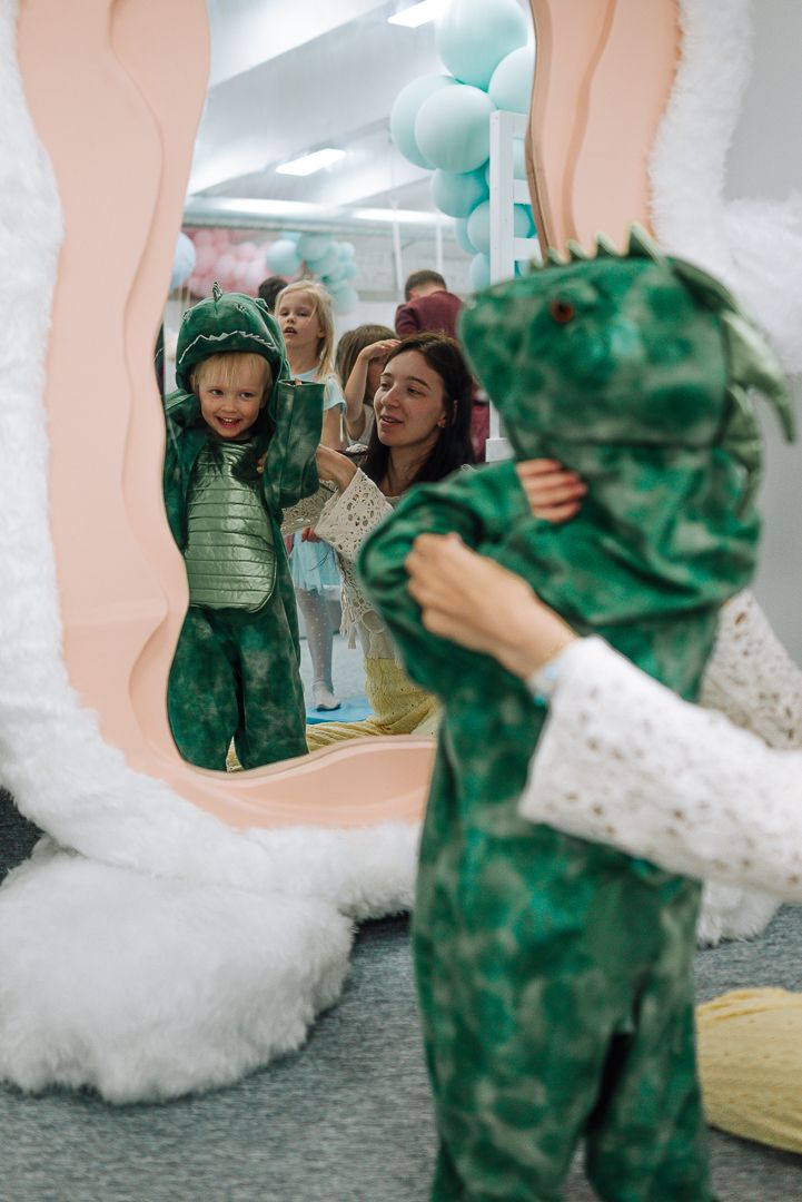 A mirror reflection shows someone in a green dinosaur costume with festive decorations and balloons in the background.
