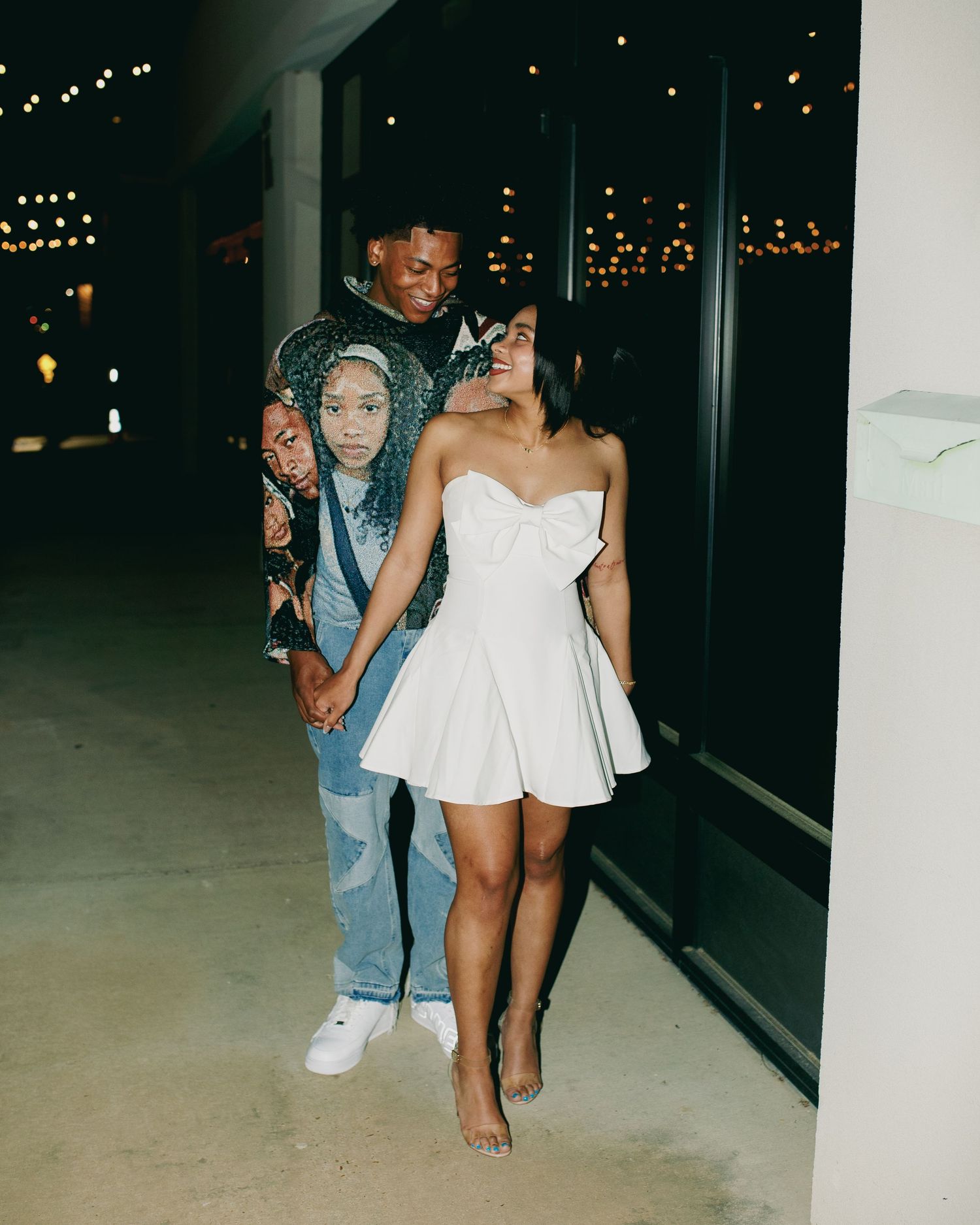 Couple posing against dark window with city lights, wearing white dress and casual denim outfit.