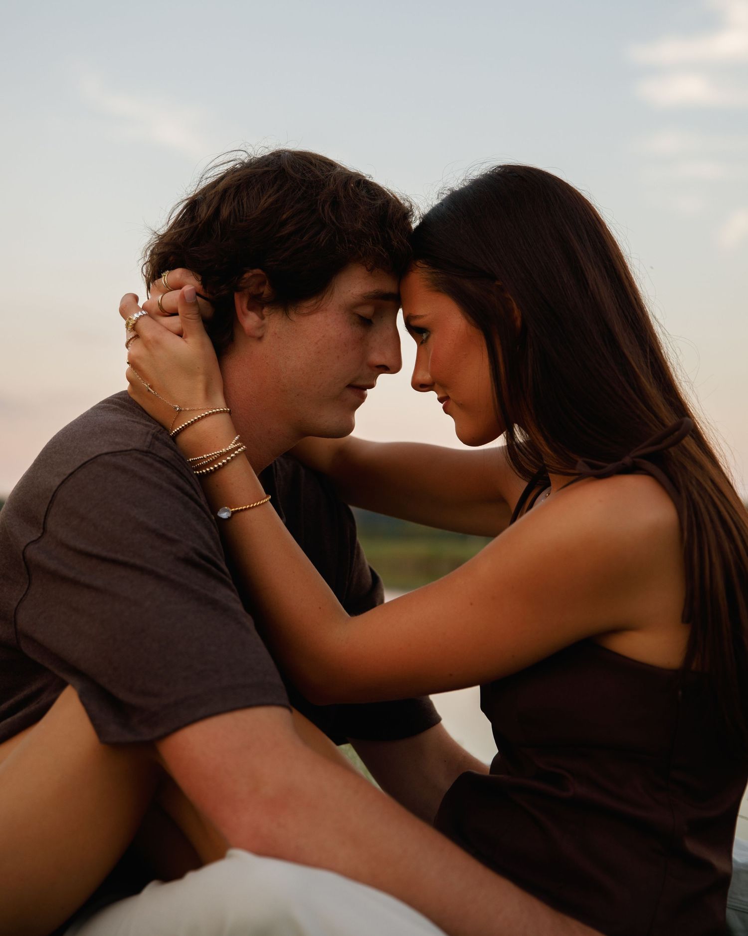 A young couple sits close together outdoors, foreheads touching in a romantic and intimate moment at dusk.