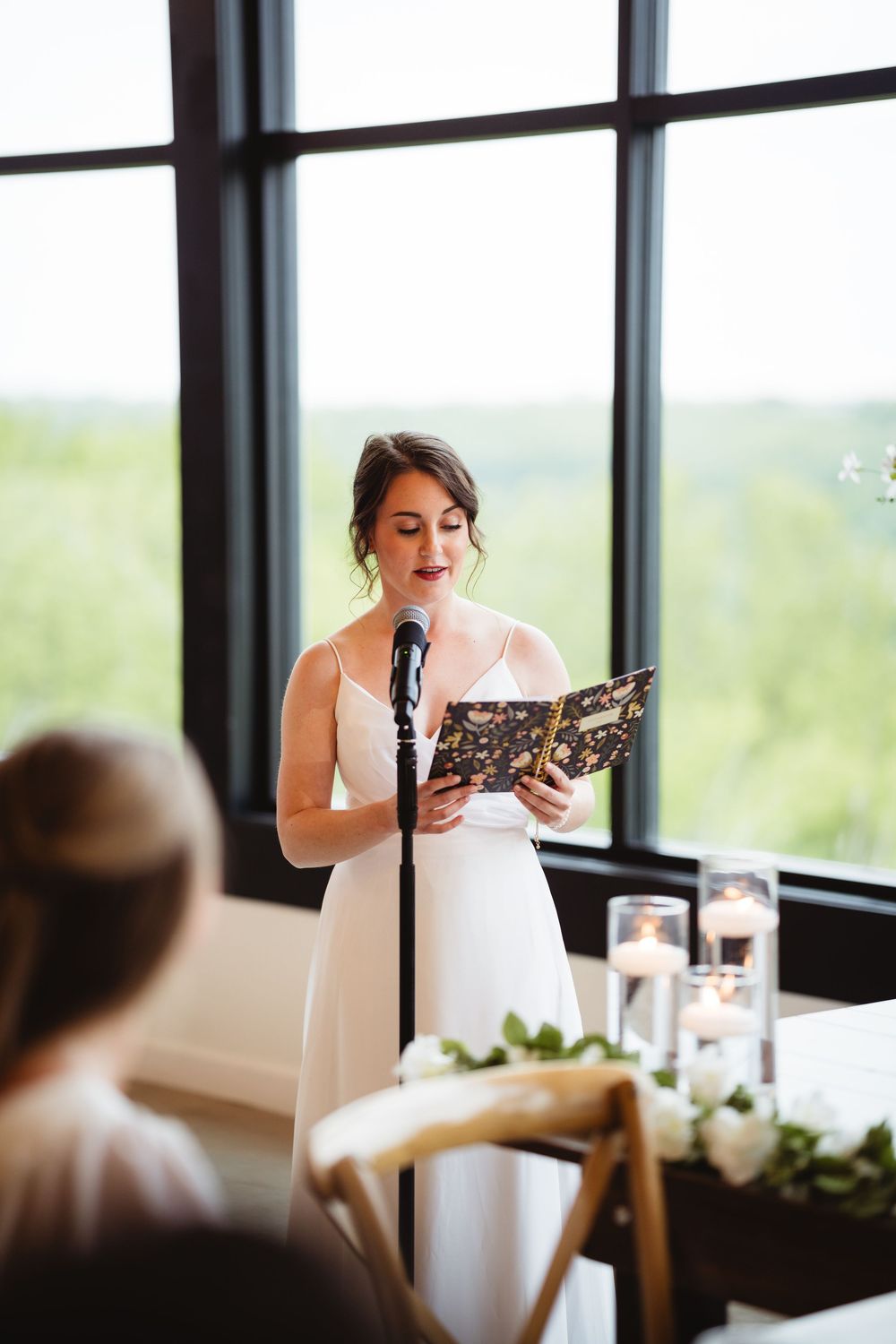 Someone in a white dress gives an emotional wedding speech at an indoor reception with large windows.