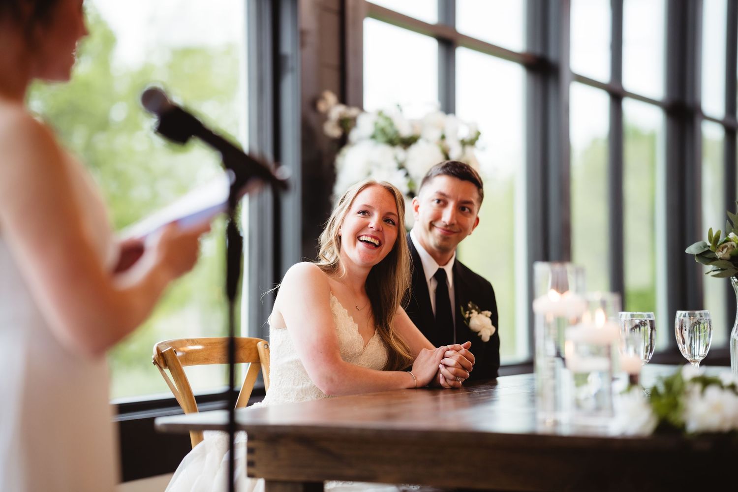 Happy couple laughs while listening to wedding toast speech at reception venue by large windows.