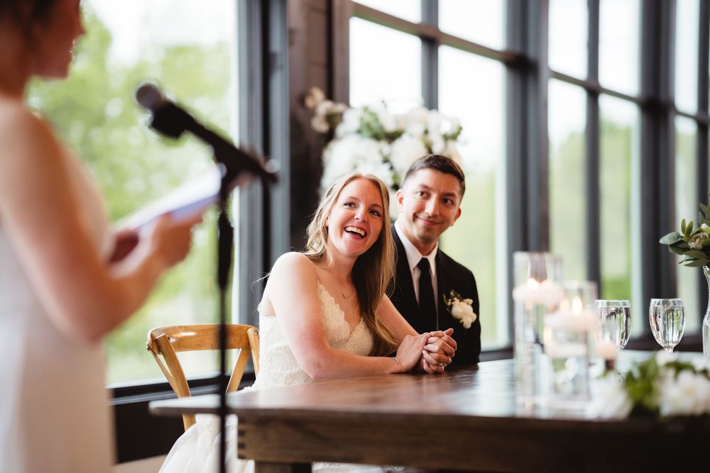 Happy couple laughs while listening to wedding toast speech at reception venue by large windows.