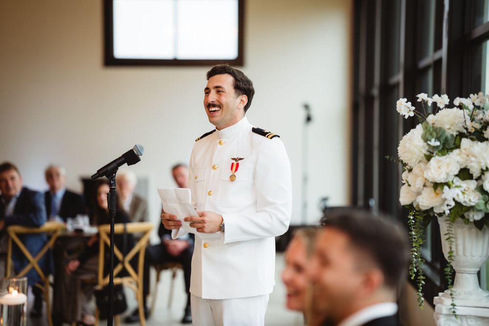 A person in a white naval uniform gives a speech at a formal event with floral decorations.