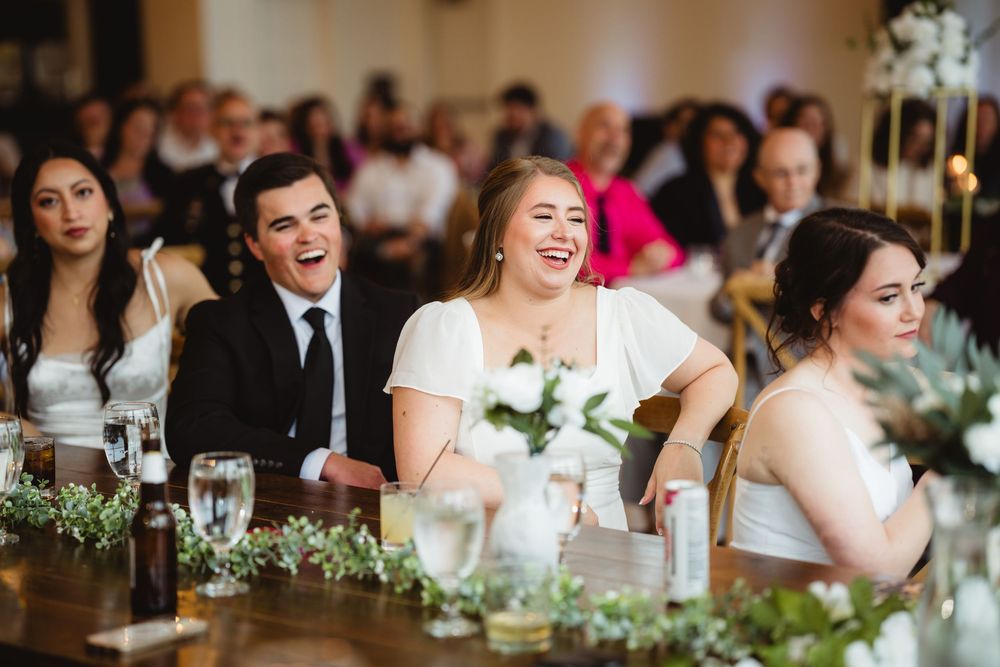 Reception guests laugh and smile while seated at a decorated wedding table with white flowers and greenery centerpieces.