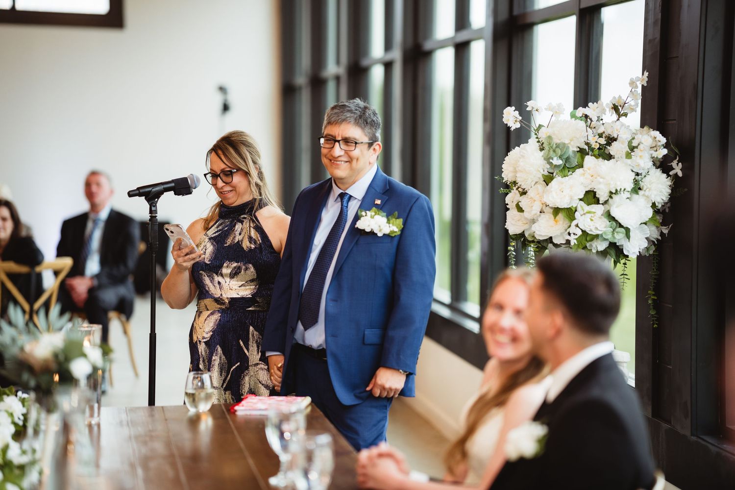 People delivering touching wedding reception speeches at an elegant venue decorated with white floral centerpieces.