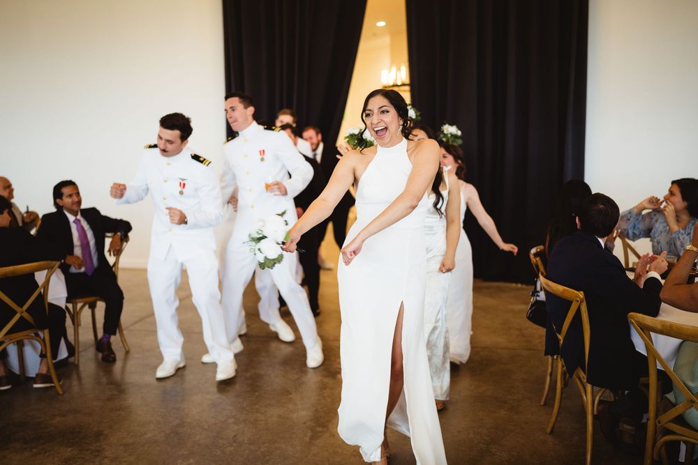 Wedding party performers in white outfits dance together during a lively celebration at an indoor venue.