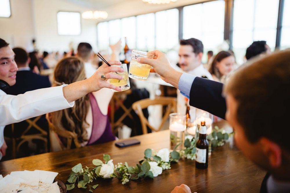 People raise their glasses in a toast across a dining table decorated with small floral centerpieces during a celebration.