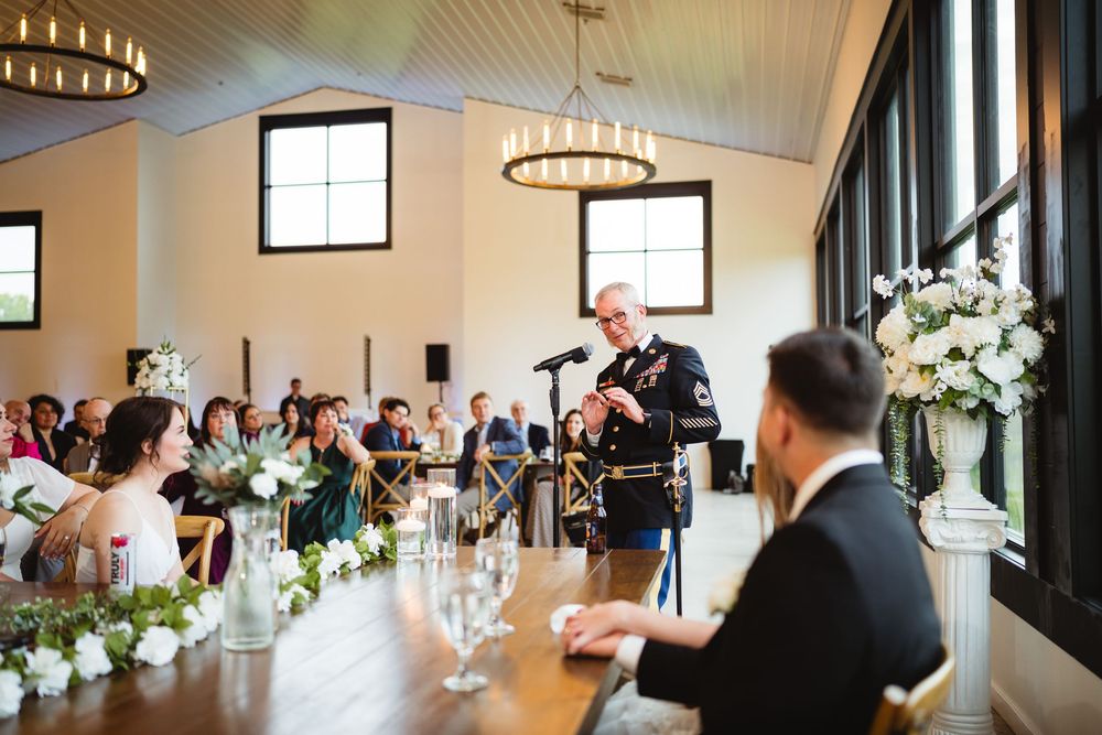 Guests listen attentively during a speech at an elegant wedding reception with floral centerpieces and modern lighting.