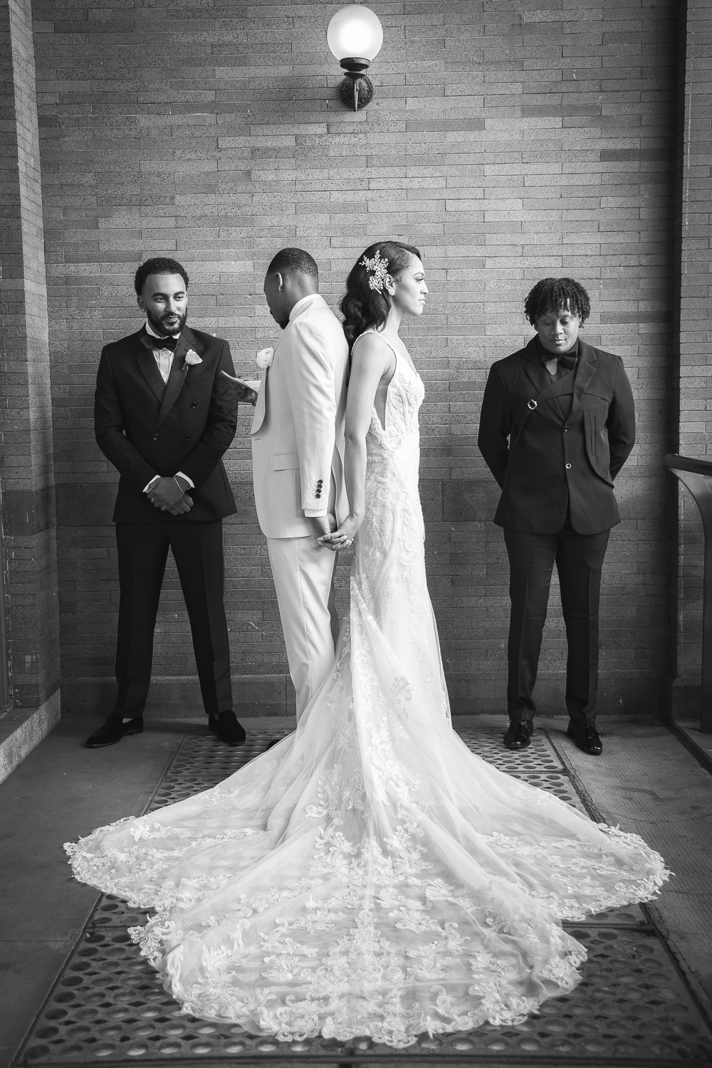 A wedding party poses in a hallway with a bride in an elegant lace gown with a dramatic train and three attendants.