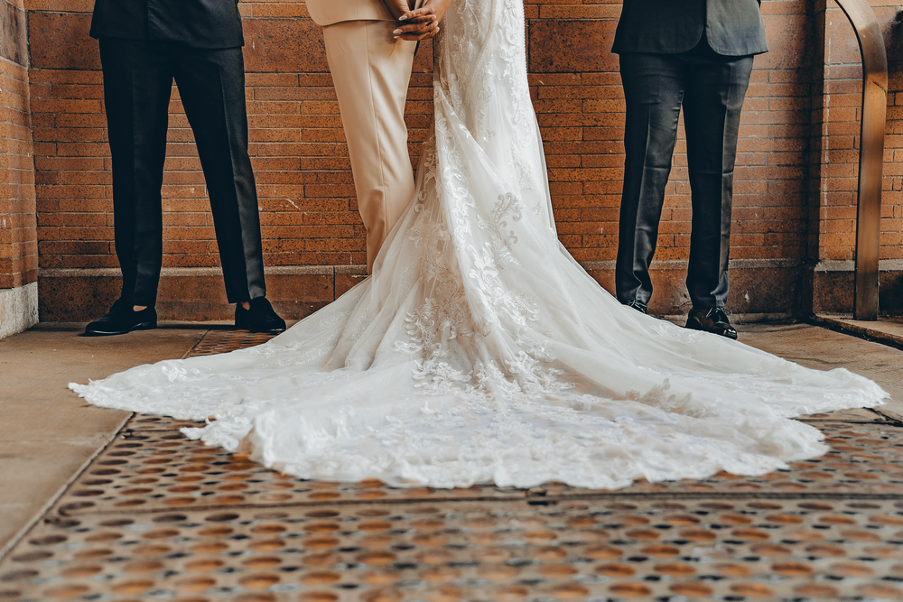 A wedding dress with intricate lace train on geometric tile flooring, flanked by suited figures.