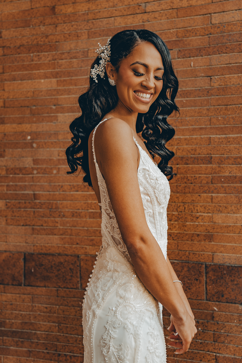 Beautiful bride in a form-fitting lace wedding gown poses against brick wall with floral hair accessory and wavy hair.