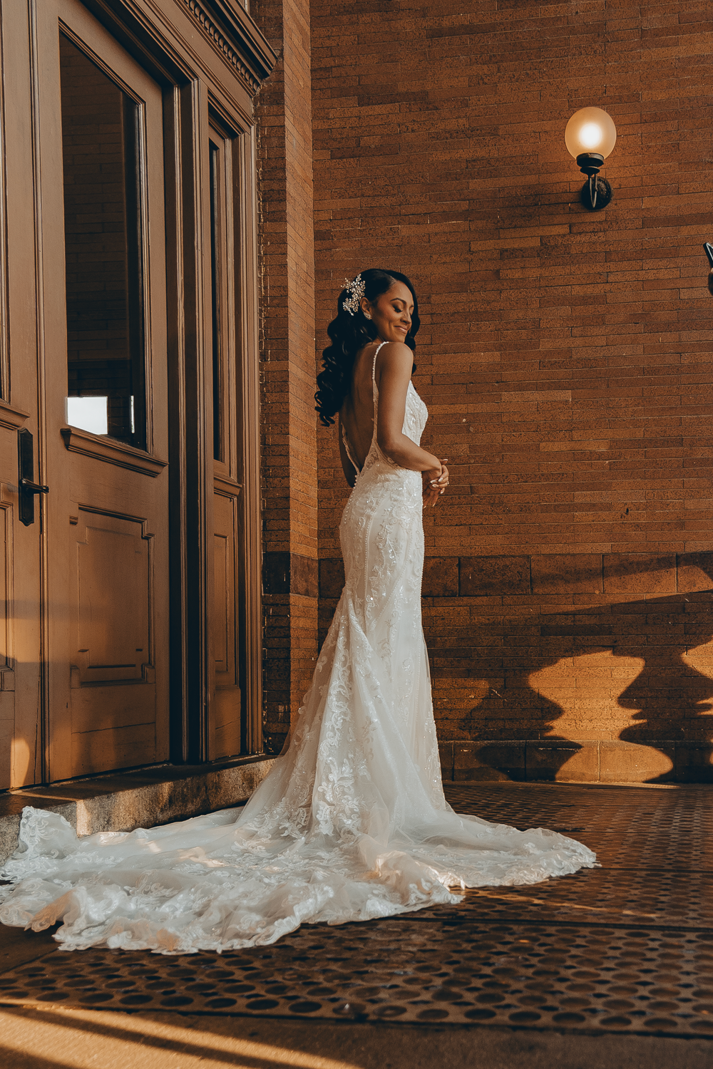 A bride in a fitted lace wedding gown with a long train poses in golden evening light against a brick wall.
