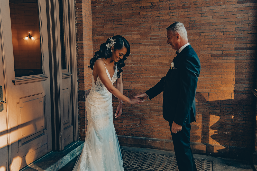 A tender moment between a bride in a fitted white lace gown and someone in a dark suit on a porch at night.