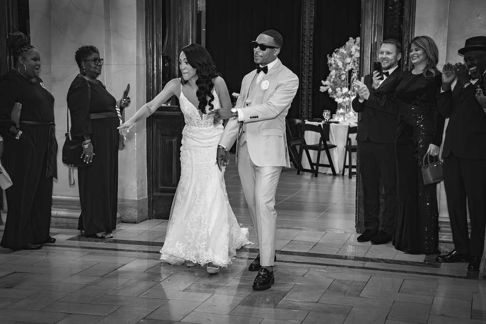 A couple in formal wedding attire dances at their reception while guests observe in black and white photography.